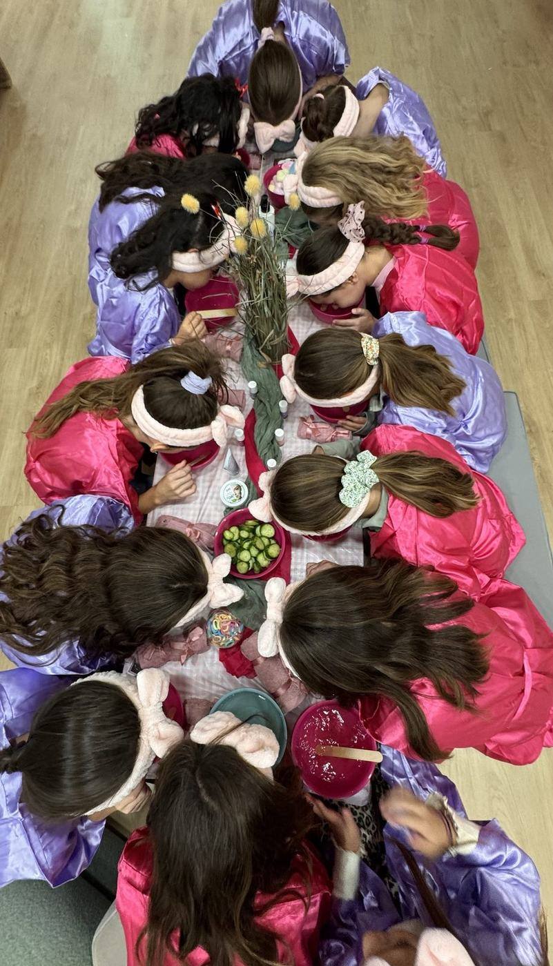 Κάτοψη παιδικού spa party: παιδιά με ρόμπες και headbands σκυμμένα πάνω από τα μπολ τους γύρω από το τραπέζι